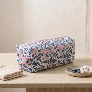 K&ouml;nnte beinhalten: Eine rechteckige, gesteppte Kulturbeutel aus Stoff mit einem blau-rosa Blumenmuster auf wei&szlig;em Grund. Die Tasche verf&uuml;gt &uuml;ber einen Rei&szlig;verschluss an der Oberseite und einen seitlichen Tragegriff aus rosa-wei&szlig; gestreiftem Stoff.