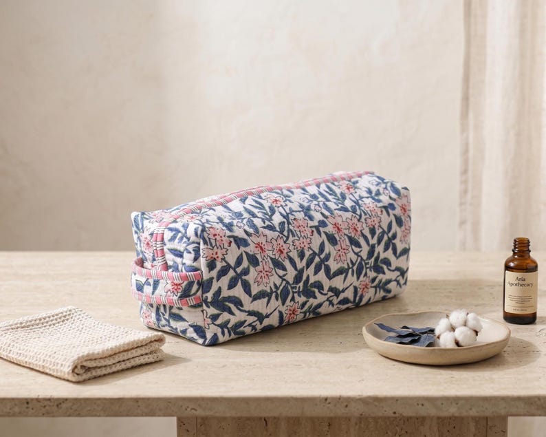 K&ouml;nnte beinhalten: Eine rechteckige, gesteppte Kulturbeutel aus Stoff mit einem blau-rosa Blumenmuster auf wei&szlig;em Grund. Die Tasche verf&uuml;gt &uuml;ber einen Rei&szlig;verschluss an der Oberseite und einen seitlichen Tragegriff aus rosa-wei&szlig; gestreiftem Stoff.