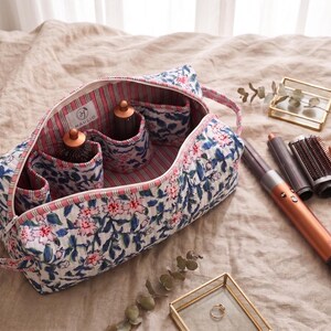 K&ouml;nnte beinhalten: Eine gesteppte Stofftasche mit blau-rosa Blumenmuster, gestreiftem Innenfutter und einem Etikett mit der Aufschrift HAULIO. Die Tasche enth&auml;lt Haarstyling-Ger&auml;te, daneben liegen weitere Aufs&auml;tze und ein Haartrockner-Stab.