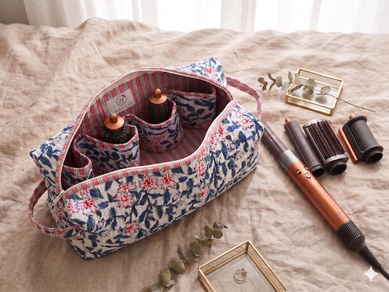 K&ouml;nnte beinhalten: Eine gesteppte Stofftasche mit blau-rosa Blumenmuster, gestreiftem Innenfutter und einem Etikett mit der Aufschrift HAULIO. Die Tasche enth&auml;lt Haarstyling-Ger&auml;te, daneben liegen weitere Aufs&auml;tze und ein Haartrockner-Stab.