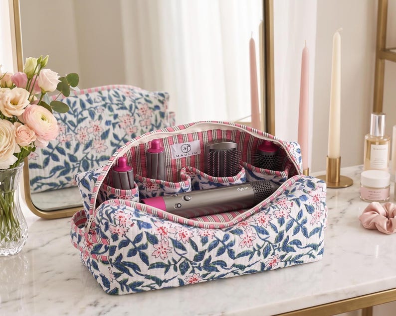K&ouml;nnte beinhalten: Eine gesteppte Aufbewahrungstasche aus Stoff mit blau-rosa Blumenmuster auf wei&szlig;em Grund. Das Innere ist mit rot-wei&szlig; gestreiftem Stoff ausgekleidet und enth&auml;lt ein graues Haarstyling-Ger&auml;t mit mehreren Aufs&auml;tzen.
