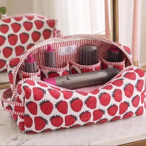 May include: A quilted fabric storage bag featuring a red strawberry pattern on a white background. The interior is lined with red and white stripes and contains multiple compartments holding a grey hair styling tool and various attachments.
