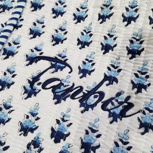 K&ouml;nnte beinhalten: Ein wei&szlig;er Steppstoff mit einem sich wiederholenden Muster aus kleinen blauen Blumenmotiven und einem dunkelblau gestreiften Rand. Der Name Tanbor ist in dunkelblauer Schreibschrift in die Mitte des Steppstoffs gestickt.
