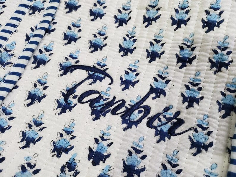K&ouml;nnte beinhalten: Ein wei&szlig;er Steppstoff mit einem sich wiederholenden Muster aus kleinen blauen Blumenmotiven und einem dunkelblau gestreiften Rand. Der Name Tanbor ist in dunkelblauer Schreibschrift in die Mitte des Steppstoffs gestickt.