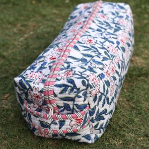 K&ouml;nnte beinhalten: Eine rechteckige, gesteppte Reisetasche mit wei&szlig;em Grund und einem sich wiederholenden Blumenmuster in Blau und Pink. Die Tasche verf&uuml;gt &uuml;ber einen Rei&szlig;verschluss oben und einen Seitengriff, beide mit einem rosa-wei&szlig; gestreiften Stoffbesatz.