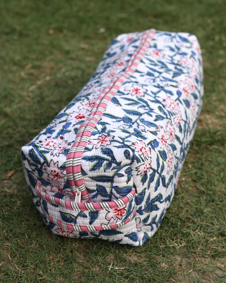 K&ouml;nnte beinhalten: Eine rechteckige, gesteppte Reisetasche mit wei&szlig;em Grund und einem sich wiederholenden Blumenmuster in Blau und Pink. Die Tasche verf&uuml;gt &uuml;ber einen Rei&szlig;verschluss oben und einen Seitengriff, beide mit einem rosa-wei&szlig; gestreiften Stoffbesatz.