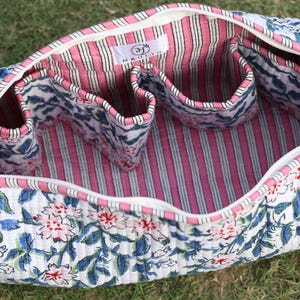 K&ouml;nnte beinhalten: Eine gesteppte Stofftasche mit wei&szlig;em Au&szlig;enmaterial und blau-rosa Blumenmuster. Das Innere ist mit rosa-wei&szlig;en vertikalen Streifen ausgekleidet und verf&uuml;gt &uuml;ber mehrere offene F&auml;cher. Ein wei&szlig;es Etikett mit der Aufschrift HAULER ist zu sehen.