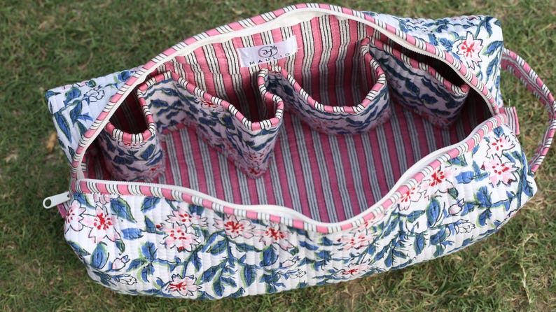 K&ouml;nnte beinhalten: Eine gesteppte Stofftasche mit wei&szlig;em Au&szlig;enmaterial und blau-rosa Blumenmuster. Das Innere ist mit rosa-wei&szlig;en vertikalen Streifen ausgekleidet und verf&uuml;gt &uuml;ber mehrere offene F&auml;cher. Ein wei&szlig;es Etikett mit der Aufschrift HAULER ist zu sehen.