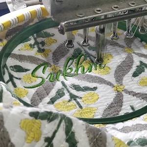K&ouml;nnte beinhalten: Ein wei&szlig;er Steppstoff mit gelbem und grauem Blumenmuster ist in einem dunkelgr&uuml;nen runden Stickrahmen eingespannt. Die Nadel einer Stickmaschine stickt den Namen Sukhman in gr&uuml;ner Schreibschrift auf den Stoff.