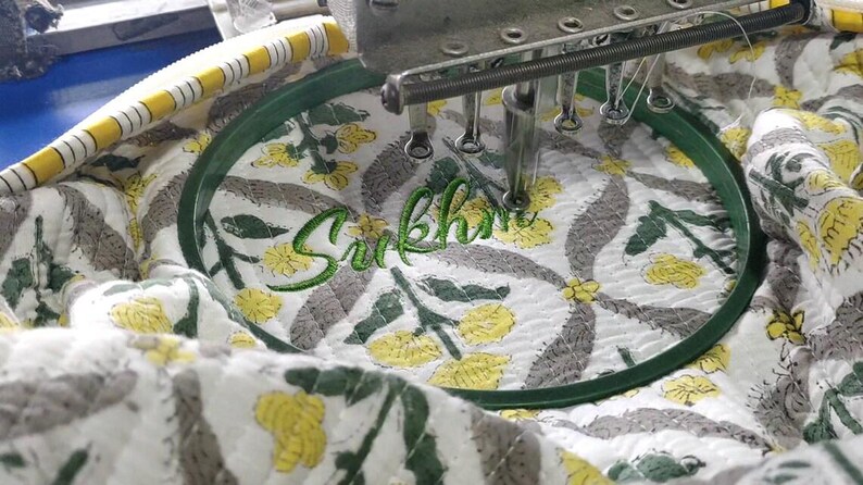 K&ouml;nnte beinhalten: Ein wei&szlig;er Steppstoff mit gelbem und grauem Blumenmuster ist in einem dunkelgr&uuml;nen runden Stickrahmen eingespannt. Die Nadel einer Stickmaschine stickt den Namen Sukhman in gr&uuml;ner Schreibschrift auf den Stoff.