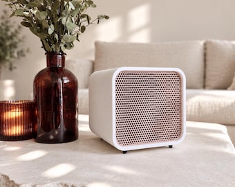 May include: A white, cube-shaped speaker with a rose gold metallic mesh front grille. The device features rounded corners and sits on small black feet, showcasing a clean, modern design with a prominent perforated speaker face.