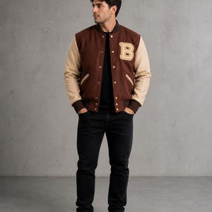 Könnte beinhalten: Braune und beige Collegejacke mit einem großen "B"-Aufnäher auf der Brust. Die Jacke hat eine Knopfleiste und beige Ärmel. Die Person trägt ein schwarzes Oberteil, eine schwarze Jeans und weiße Turnschuhe.