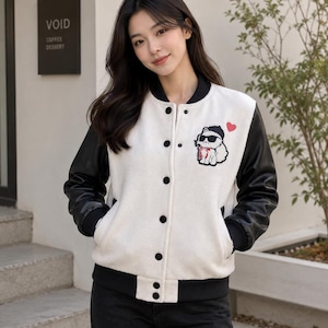 Könnte beinhalten: Weiß-schwarze Collegejacke mit einem Cartoon-Katzen-Patch, die eine Sonnenbrille und eine rote Fliege trägt. Die Jacke hat schwarze Ärmel, einen schwarzen Kragen und schwarze Knöpfe. Ein kleines rotes Herz befindet sich neben dem Katzen-Patch.