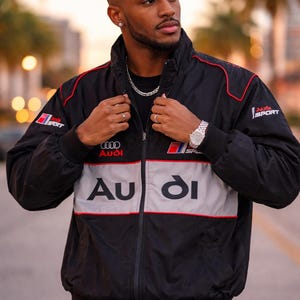 May include: Black zip-up jacket with red trim and the Audi logo. The jacket has a gray stripe across the chest with the Audi logo and the words "Audi Sport". The jacket is worn by a person.