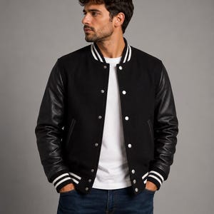 Könnte beinhalten: Schwarze Collegejacke mit Lederärmeln, weißen Streifen an den Bündchen und am Kragen sowie weißen Druckknöpfen. Die Jacke wird über einem weißen T-Shirt und einer blauen Jeans getragen. Ein klassisches, stilvolles Oberbekleidungsstück.