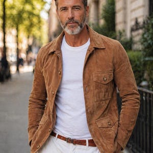 Könnte beinhalten: Braune Wildlederjacke mit vier Fronttaschen und Knopfverschlüssen. Die Jacke wird über einem weißen T-Shirt getragen und mit einer weißen Hose und einem braunen Ledergürtel kombiniert. Das Outfit hat einen klassischen, lässigen Stil.