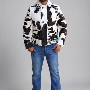 Könnte beinhalten: Kuhfelljacke mit schwarz-weißem Muster. Die Jacke hat eine Knopfleiste, zwei Taschen und einen Kragen. Die Person trägt eine blaue Jeans und weiße Turnschuhe. Die Jacke ist ein Statement-Piece.