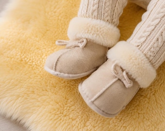 May include: A pair of beige suede booties with cream-coloured faux fur trim and small decorative bows, worn with cream-coloured cable-knit leggings. The booties feature a soft, rounded toe design and a plush interior lining visible at the ankle cuffs.