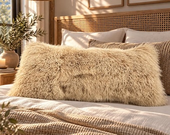 May include: A rectangular throw pillow featuring a long, shaggy, cream-coloured faux fur texture. The pillow is positioned horizontally on a bed covered with a light-coloured textured blanket, with a woven headboard visible in the background.