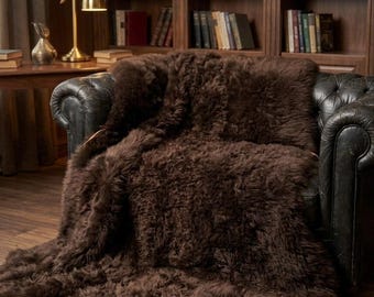 May include: A dark brown sheepskin rug with long, thick, and dense fur. The rug is draped over a dark leather armchair, covering the seat and backrest, with the lower portion extending down to the floor in a natural, irregular shape.