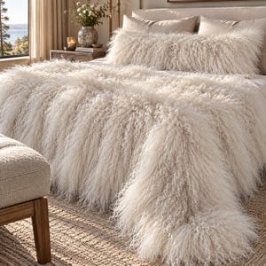 May include: A long, cream-colored throw blanket with a thick, shaggy, curly texture draped over a bed. A matching long, rectangular bolster pillow with the same shaggy, curly cream-colored texture is positioned at the head of the bed.