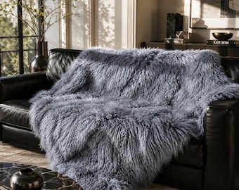 May include: A long-haired, shaggy throw blanket in a cool slate blue color. The blanket features a dense, textured pile with a wavy, crimped appearance that drapes over the surface of a black leather sofa, covering the seat and back cushions.