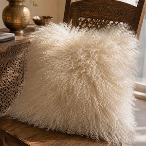 May include: A square throw pillow covered in long, curly, off-white sheepskin fur. The texture is dense and shaggy, featuring tight, spiraled curls that cover the entire front surface of the cushion, creating a voluminous and soft appearance.