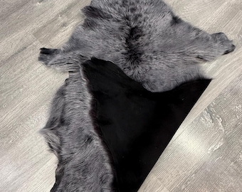 May include: A pelt with long, thick, grey and black variegated fur on one side and a smooth, solid black surface on the other. The hide is folded over to reveal both the textured, multi-tonal fur and the plain, dark underside of the material.