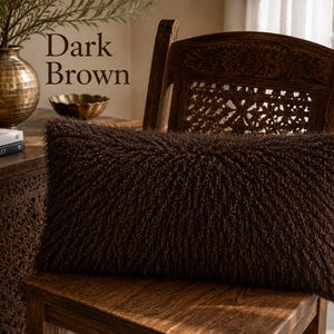 May include: A rectangular dark brown throw pillow with a shaggy, textured surface. The pillow features long, curly fibers covering the entire front face, creating a dense and uniform appearance. The text Dark Brown is visible in the image.