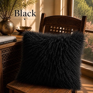 May include: A square throw pillow with a long, curly black faux fur texture. The pillow is positioned on a wooden chair. The word Black is printed in a serif font above the pillow, accompanied by a small diamond icon and a horizontal line.