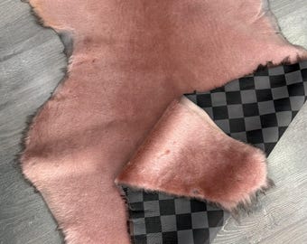May include: A soft, dusty pink animal hide rug with a dense, plush texture. One corner of the hide is folded over to reveal a contrasting underside featuring a black and grey checkered pattern, showcasing the dual-sided design of the material.
