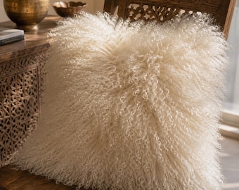 May include: A square throw pillow featuring long, curly, off-white Mongolian lamb fur. The texture is dense and shaggy, covering the entire front surface of the cushion, creating a voluminous and soft appearance with a natural, cream-colored hue.