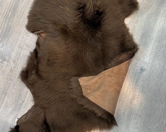 May include: A dark brown sheepskin rug with a thick, dense fur texture. The rug features an irregular, organic shape with a smooth, tan-coloured leather underside partially folded over to reveal the natural hide backing of the pelt.