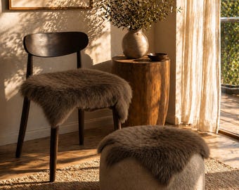 May include: A dark wood chair with a brown faux fur seat cover, a cylindrical wood side table holding a ceramic vase with branches, and a round, textured beige ottoman topped with a matching brown faux fur cover.