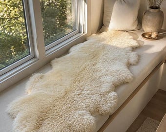 May include: A long, cream-coloured sheepskin rug with a thick, shaggy texture and irregular, natural edges. The rug is laid out flat, showcasing its dense, curly wool fibres and soft, off-white hue, providing a plush and tactile surface appearance.