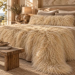 May include: A cream-colored, long-pile shaggy throw blanket draped over a bed, accompanied by a matching long rectangular bolster pillow. The texture is dense and curly, covering the surface of the bed in a soft, layered appearance.
