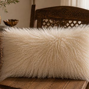 May include: A rectangular throw pillow featuring long, curly, off-white faux fur fibers that create a dense, textured appearance. The pillow has a soft, shaggy surface and is positioned on a wooden chair, showcasing its fluffy, voluminous design.