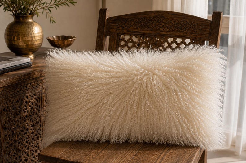May include: A rectangular throw pillow featuring long, curly, off-white faux fur fibers that create a dense, textured appearance. The pillow has a soft, shaggy surface and is positioned on a wooden chair, showcasing its fluffy, voluminous design.