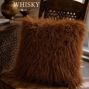 May include: A square throw pillow with long, curly, rust-brown fur texture resting on a dark wooden chair. The word WHISKY is printed in white capital letters above the pillow, accompanied by a small decorative diamond-shaped graphic element.