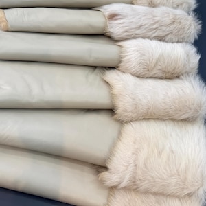 Puede incluir: Una pila de piezas de cuero beige claro plegadas, cada una con un borde de piel color crema grueso y texturizado en un lado. Las superficies de cuero liso están dispuestas horizontalmente, mostrando el contraste entre la piel mate y el pelaje denso.