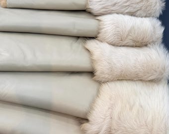 May include: A stack of folded light beige leather pieces, each featuring a thick, textured cream-coloured fur trim along one edge. The smooth leather surfaces are layered horizontally, showcasing the contrast between the matte hide and the soft, dense fur.