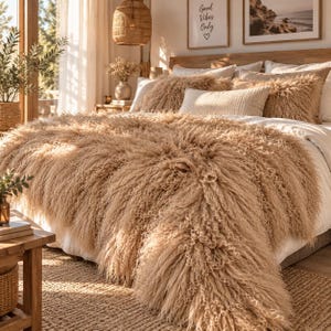 May include: A beige, long-pile faux fur throw blanket draped over a bed with white linens. The bed features several decorative pillows, including two shaggy textured pillows and one rectangular cream-colored pillow, all in a neutral color palette.