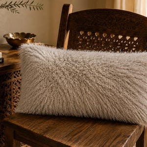 May include: A rectangular throw pillow with a long, shaggy, off-white faux fur texture. The pillow has a dense, fibrous appearance with a soft, fluffy pile that covers the entire front surface, creating a uniform and highly textured look.