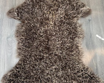 May include: A brown and grey sheepskin rug featuring long, dense, and curly wool fibres. The pelt has an irregular, organic shape with a thick, textured pile that displays a natural gradient of earthy tones throughout the entire surface area.