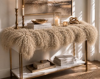 May include: A console table with a gold metal frame and white marble shelves, draped with a beige curly faux fur throw. The top shelf holds two brass candlesticks, a gold bowl, two books titled Kinfolk and Live Beautifully, a glass candle lantern, and a gold knot sculpture. The bottom shelf holds a rectangular box and three stacked books.