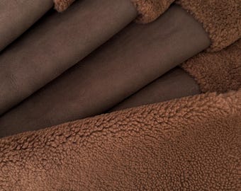 May include: A folded piece of fabric featuring a smooth, dark brown suede-like side and a contrasting thick, textured, medium brown faux shearling side. The material is layered in multiple folds, showcasing the dual-sided texture and colour.