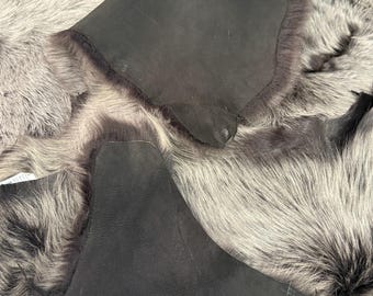 May include: A collection of sheepskin pelts featuring a dual-sided texture. One side displays a dense, long-haired coat in shades of grey and silver, while the reverse side shows a smooth, matte dark brown leather surface with irregular edges.