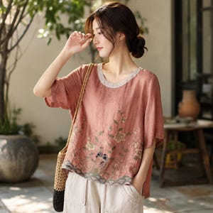 May include: A dusty rose short-sleeved blouse featuring a round neckline with a light grey trim. The front of the garment is decorated with an embroidered floral pattern in muted tones, including a small blue bird detail near the bottom hem.