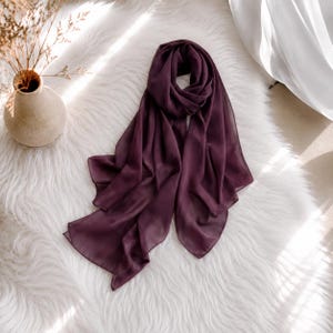 May include: A deep plum-colored rectangular scarf made of a lightweight, semi-sheer fabric. The scarf features a smooth, flowing texture with soft folds and drapes, showcasing a solid, consistent dark purple hue throughout the entire piece.