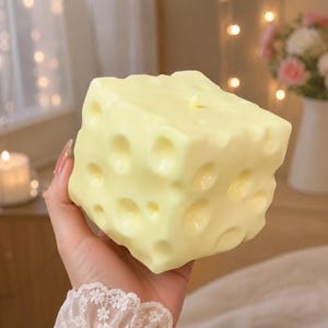 May include: A pale yellow, cube-shaped candle designed to resemble a block of Swiss cheese. The surface features multiple circular indentations of varying sizes across its sides and top, with a small, centered wick protruding from the top surface.